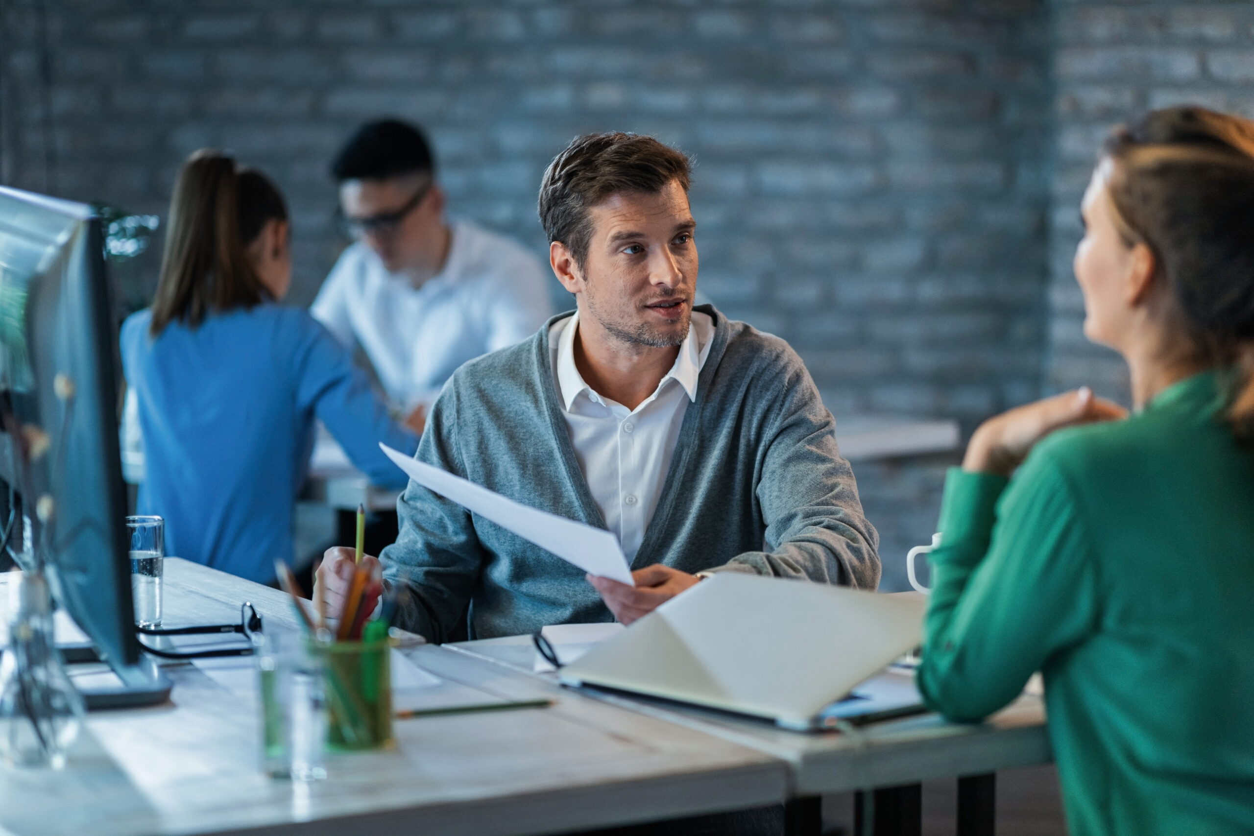 male-entrepreneur-discussing-about-business-reports-with-female-colleague-while-working-office-desk-there-are-people-background